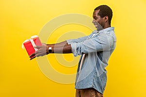 Take this present! Side view of generous man in denim casual shirt holding out red gift box. indoor studio shot isolated on yellow