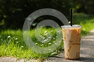 Takeaway plastic cup with cold coffee drink and straw near green grass outdoors, space for text