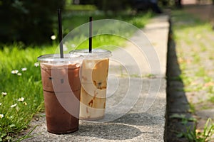 Takeaway plastic cups with cold coffee drinks near green grass outdoors, space for text