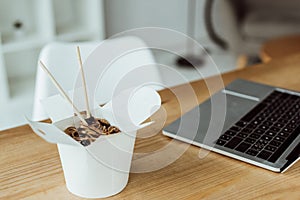 takeout box with chinese noodles and chopsticks on table with laptop