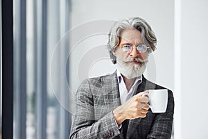 Takes a break and drinks coffee. Senior stylish modern man with grey hair and beard indoors