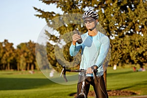 Taking a break. Young athletic man in sportswear and protective helmet standing with his bike in park, drinking water