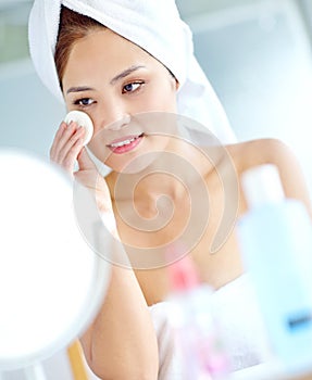 Taking care of her complexion. An attactive young Asian woman applying moisturizer with a towel on her head.