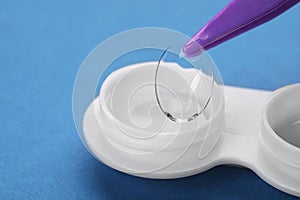 Taking contact lens from case with tweezers on blue background, closeup