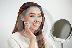 Taking off her make-up. Beautiful cheerful young woman using cotton disk and looking at her reflection in mirror with smile while