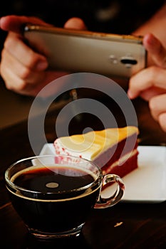 Taking photo with black coffee and red velvet cake on dark wooden table with smartphone with man hand. for up to social network.