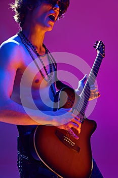 Talented and artistic young man, shirtless musician playing electric guitar against pink background in neon light