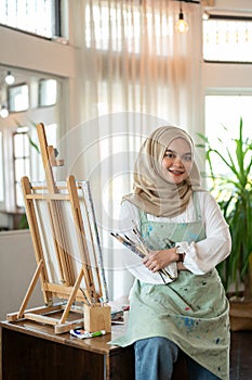 A talented Asian-Muslim female artist is standing in her studio with painting brushes in her hand