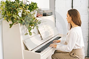 Talented attractive young woman musician performer playing gentle music on white classical piano in light home studio.