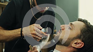 Talented bearded barber is trimming the beard of his client in a black cutting hair cape in the barbershop. He is using