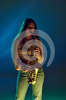 Talented, beautiful, young woman, saxophonist in gold jacket plays in neon light against gradient background.