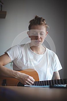 Talented boy playing a wooden acoustic guitar in a lighted room. Blonde Man aged 20-29 strums guitar and sings from hymn book on