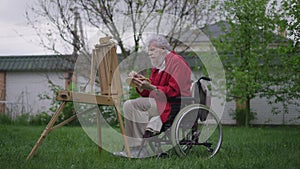 Talented creative disabled old man painting on easel in spring summer garden on backyard in slow motion. Side view wide