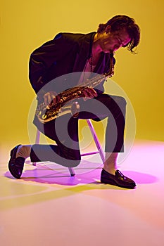 Talented jazz man, virtuoso playing saxophone sitting on chair in neon light against vibrant yellow studio background.