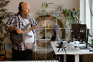 Mature composer plays bas guitar recording track at workplace in home studio