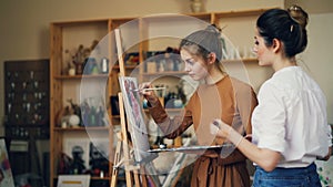 Talented student of art school is painting picture with her teacher working together in studio full of artworks and