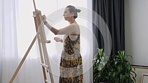 Talented young female artist standing near easel with brush in hand and painting on canvas. Beautiful woman in art home studio is