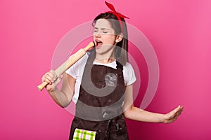 Talented young woman holds rolling pin in her hand pretented its microphone, opens mouth while singing. Energetic cute lady wears photo