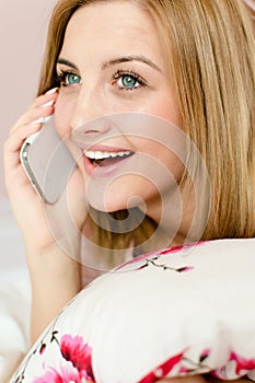 Talking on the mobile cell phone happy smiling charming young blond woman lying in bed holding pillow