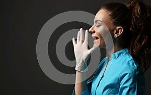 Talking or screaming young woman doctor in blue medical gown and latex gloves holds hand with fingers spread at face