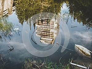 A tall abandoned building surrounded by a forest is reflected in the green water of a pond overgrown with grass and