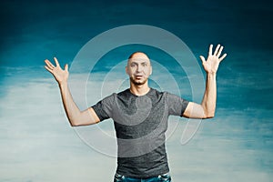 A tall bald man in a gray T-shirt shouts something emotionally, raising his hands on a blue background.