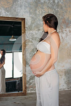 Tall beautiful pregnant girl young woman with long curly hair standing in front of the mirror in loft studio