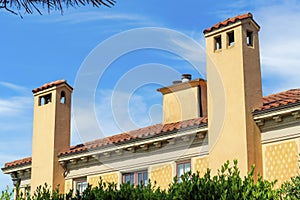 Tall beige stucco chimney pipes on house or home exterior with backyard trees and foliage in shade with building in sun