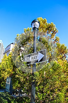 A tall black light post with a black and white `One Way` sign surrounded by lush green trees with blue sky in downtown Atlanta