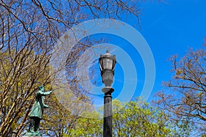 A tall black metal lamp post surrounded by bare winter trees, lush green trees and a statue with blue sky and clouds