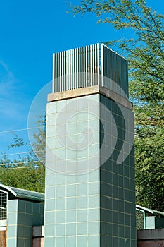 Tall cement chimney decorated with green tiles and metal vent on top in late afternoon sun on building in public area