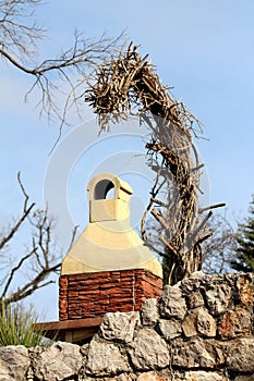 Tall dry tree without branches leaning above homemade brick barbeque in front of traditional stone wall with clear blue sky in