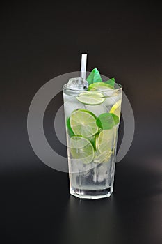 Tall faceted glass of refreshing lemonade with ice and mint on a black background, next to pieces of ripe lime