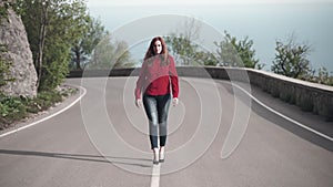 A tall girl in a red shirt and red hair is walking along the dividing line along the center of the road.