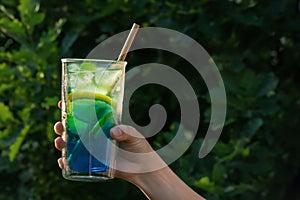 Tall glass with a blue lagoon cocktail in a female hand against the backdrop of greenery in the yard. Selective focus