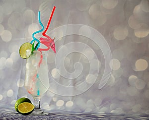 Tall glass of clear fruit nectar with ice and straws, decorated with a slice of lime on a gray glittering background