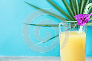 Tall Glass with Freshly Pressed Pineapple Orange Coconut Juice Straw and Small Flower. Round Palm Tree Leaf on Blue Background