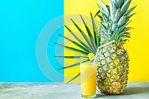 Tall Glass with Freshly Pressed Pineapple Orange Coconut Juice Straw Flower. Round Palm Leaf on Duotone Blue Yellow Background