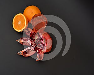 A tall glass of fruit juice on a black background, next to orange halves and a broken ripe pomegranate fruit with seeds