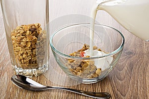 Tall glass with fruit muesli, yogurt poured on granola in bowl, spoon on wooden table
