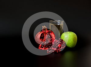 A tall glass of mixed fruit juices on a black background, next to a ripe green apple and a broken pomegranate fruit with seeds