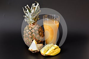 A tall glass of pineapple juice with mango and pieces of ripe fruit on a black background
