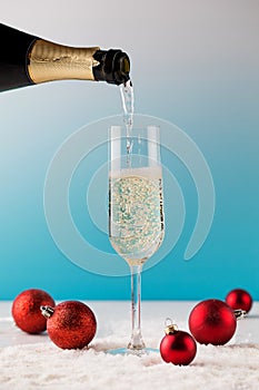 A tall glass is pouring champagne from a bottle on artificial snow with Christmas balls on a blue background.