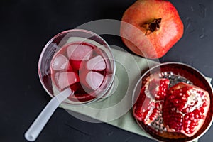 A tall glass of red juice with ice and straws on a gray background, next to a ripe pomegranate fruit