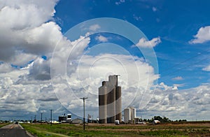 Tall grain elevators in the midwestern United States sorrounded by trucks and equipment and powerlines beside a highway all under