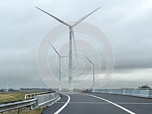 Tall green energy wind mills or turbines near a highway under a cloudy sky