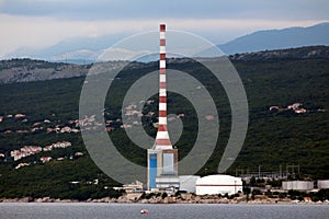 Tall industrial chimney next to large industrial oil refinery tankers surrounded with dense forest and family houses next to calm