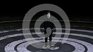 Tall man in black clothes standing in the meddle of amphitheater. Stock clip. Asphalt colored in black circles.