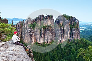 Tall man with red baseball cap sit on cliff and watch to mountains. Dark silhouette of rocks.