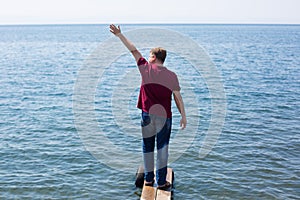 A tall man standing on a narrow plank by the water waves his hand seeing off a friend